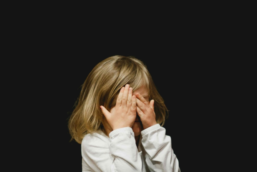 A child is covering their face with their hands against a dark background.